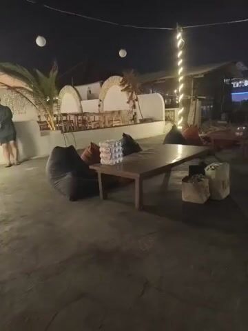 The video shows an outdoor seating area at night. In the foreground, there is a wooden table surrounded by beanbag chairs in dark colors. A person in a dark shirt and shorts stands near a white wall on the left side of the frame, leaning against it. Further back, there is a building with arched openings and some seating. String lights are visible, decorating poles and extending across the space. In the background, there are more structures and lights suggesting a town or resort area. There are also decorative white spherical lanterns hanging overhead. The overall impression is of a relaxed, dimly lit evening gathering space.
