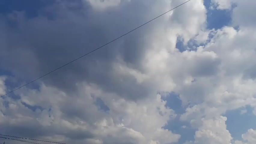 This is a still image taken outdoors, likely during the day. The primary subject is the sky, filled with a mix of white, puffy cumulus clouds and larger, darker grey clouds. The clouds suggest dynamic weather, possibly with some sun breaking through in patches. A thin, dark line, likely a power line or telephone wire, stretches diagonally across the upper portion of the frame. The setting appears to be an open area, with no prominent structures or landscape features visible, except for a hint of dark foliage or a wire at the bottom left corner, suggesting the presence of poles or trees at a lower elevation. The time of day is likely afternoon, given the brightness and the contrast of the clouds. The weather appears to be partly cloudy, possibly transitioning between sun and potential rain. There is no visible text in the image. The overall mood evoked is one of natural beauty and the vastness of the sky.