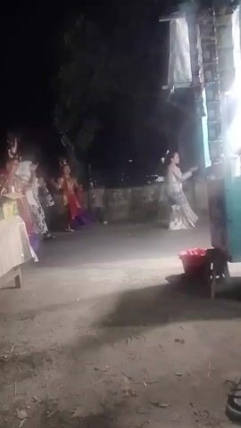 A group of at least five women in traditional clothing are dancing in an outdoor area at night. The women are wearing brightly colored outfits, including what appears to be sarongs, blouses, and headpieces. They are moving in a coordinated fashion, with some holding their arms up and others with hands clasped. The environment appears to be a street or pathway, with a building or structure on the right side of the frame that has lights and merchandise displayed on it. There are trees and foliage in the background, and the ground is uneven and dusty. The lighting is dim, with artificial lights illuminating the dancers.