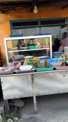 The video shows an outdoor food stall. A glass display case sits on top of a table, filled with various food items. The stall is set up against a wall with an orange and blue facade. To the right of the display case, a person is partially visible behind the counter, wearing a headscarf. The table displays more food items in plastic containers and wrappers. The ground in front of the stall appears to be dirt and gravel. A single lightbulb hangs from the ceiling above the stall.