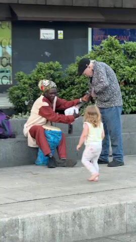The video shows three people on a city sidewalk. A Black man wearing a patterned headwrap and maroon and cream clothing sits on a low concrete ledge playing a drum. A small barefoot girl in patterned pajamas stands in front of him, looking at the drum. An older White man wearing a black cap, floral-print shirt, and jeans stands to the right of the girl, handing the seated man a crumpled white object. Behind the figures are green bushes, and the side of a building with signs can be seen in the background.