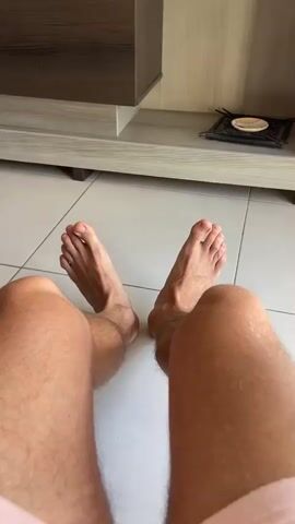The video shows a first-person perspective of a person's legs and feet. The person is sitting on a tiled floor with their legs bent at the knees. The feet are bare and appear to be moving slightly. In the background, there is a piece of furniture, possibly a cabinet or shelf, and a wall. The lighting suggests an indoor environment.