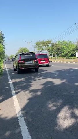 This video captures a street scene in Ketapang, Indonesia, on a bright, sunny day. The focus is on traffic moving along a paved road. In the foreground, a person wearing a green Gojek jacket and helmet rides a white scooter. They are followed by several other individuals on motorcycles, also navigating the road.

Further down the road, a few cars are visible. One is a dark-colored SUV or minivan, and another is a bright red sedan. A large yellow truck, possibly a tanker, is also in motion. The road is bordered by lush green trees and vegetation on the left and right sides. Power lines and streetlights suggest an urban or semi-urban environment. The time of day appears to be daytime, with clear blue skies and distinct shadows cast on the road, indicating the sun is high. The overall impression is one of everyday commute or delivery activity in a tropical setting.

Visible text includes "Gojek" on the driver's jacket and the license plates on some of the vehicles, though the text is not clearly legible in all instances. The presence of Gojek, a popular ride-hailing and delivery service in Southeast Asia, strongly suggests the activity involves transportation or delivery services.