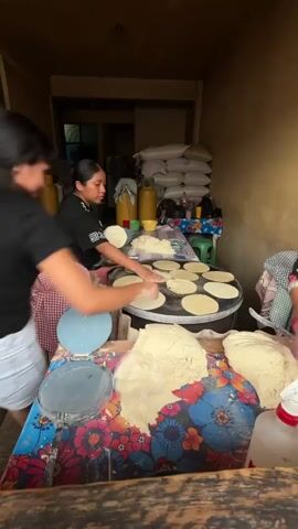The video shows a woman in a black shirt and white pants standing behind a large, round cooking surface with several tortillas on it. She is working with dough, pressing it into flat circles and placing them on the hot surface. To her left, another person's hands are visible, and a red and white checkered cloth is draped over a basket. In the foreground, a table is covered with a colorful, floral-patterned cloth, and there are mounds of dough and tortilla-making tools. A bottle with a red cap is also on the table. In the background, there are shelves stacked with sacks, a yellow container, and other items. The environment appears to be an indoor cooking area, possibly a small business or a home kitchen.