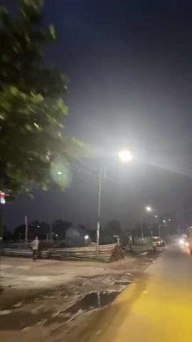 A nighttime video filmed from a moving vehicle captures a street scene with a bright streetlamp in the foreground. On the left side of the road, there are piles of logs and small, rustic shacks. A man in a light-colored shirt and a lungi walks along the muddy shoulder. In the distance, a yellow truck is parked, and a person appears to be interacting with it. Several vehicles with headlights on are driving along the wet road. The sky is dark, and the bright streetlights illuminate the immediate surroundings.