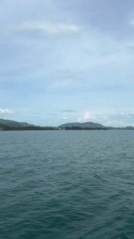 The video shows a wide view of the ocean with a distant shoreline under a partly cloudy sky. The water is a deep blue-green with small waves rippling across the surface. The shoreline is lined with green vegetation and hills, with a few buildings visible in the distance. The sky is a pale blue with scattered white clouds. The camera appears to be stationary and mounted on a boat, looking out towards the horizon.