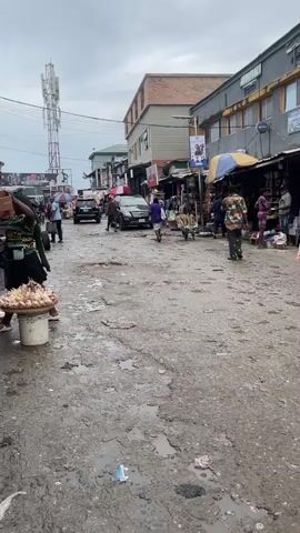 The video shows a street scene that appears to be a market. The ground is uneven and wet, with puddles and debris scattered around. On either side of the street, there are market stalls with various goods displayed, covered by umbrellas and awnings. People are walking through the market, some carrying items. A few vehicles are present, including cars and what looks like a cart being pushed. In the background, there are buildings, one of which has a tall cell tower nearby. The sky is overcast.