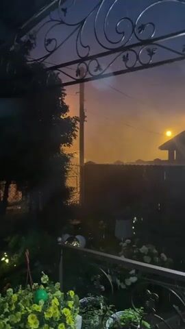 The video shows a view from a covered porch or patio looking out at a yard and beyond. It is dark, and raining, with visible streaks of rain in the air. In the background, there is a bright orange glow illuminating the sky, possibly from streetlights or a distant fire. There are trees and fences visible, as well as some potted plants on the porch. The overall impression is that of a stormy evening.