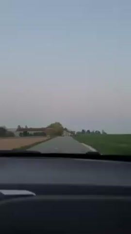 From the interior of a car moving along a road, the camera shows a rural landscape. The sky is a pale, muted blue, suggesting dawn or dusk. On the left, a long, low building complex is visible in the distance. Across the road on the right, a field of green stretches towards the horizon. As the car moves, the camera briefly catches a glimpse of a tall, white tower standing on a grassy incline. The perspective is from the driver's seat, with the dashboard visible at the bottom of the frame.
