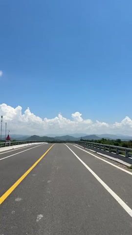 A first-person perspective video shows a clear, bright blue sky with scattered white clouds. A paved road with distinct yellow and white lane markings stretches towards a distant, hazy horizon. On either side of the road, metal guardrails are visible, and beyond them, rolling green hills dotted with trees form the landscape. The road appears to be on an elevated structure, possibly a bridge, given the presence of the guardrails and the expanse of the road. There are no people or vehicles visible in the frame. The lighting suggests it is daytime, likely late morning or early afternoon.