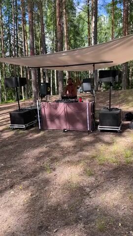 A DJ is performing at a setup in a forest. They are wearing an orange shirt and appear to be mixing music. The DJ booth has a screen and various audio equipment, including speakers mounted on stands. A canopy covers the DJ booth and equipment, providing shade. The ground is covered in pine needles and dirt.