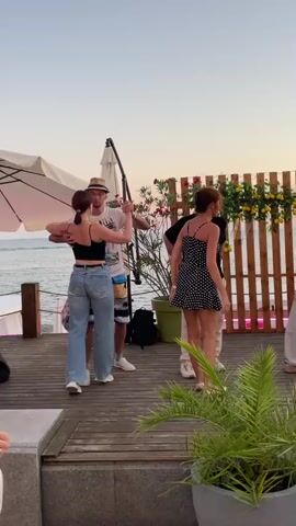 A group of people are dancing on a wooden deck near a body of water, possibly the ocean, during what appears to be sunset. The sky is a pale orange and blue. Two couples are dancing in the middle ground. A woman in a black and white polka dot dress is dancing with a man in a dark t-shirt and light-colored shorts. Another couple, a man in a white t-shirt and hat and a woman in a black tank top and jeans, are dancing in the foreground. In the background, there is a wooden fence decorated with yellow flowers and greenery. A large umbrella is visible to the left. Several potted plants are also present. The people in the foreground are facing away from the camera, looking towards the dancers. One woman with long, dark red hair is partially visible on the left side of the frame, wearing a white shirt. Another person with short, dark hair is visible in the lower left corner, wearing a patterned shirt. A man with gray hair is dancing with a woman in a white t-shirt and light-colored pants. The overall atmosphere is relaxed and festive.