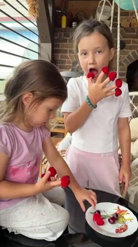 Two young girls are sitting indoors near a fireplace. The girl on the left, wearing a pink t-shirt, is seated and holding raspberries in her hands, looking down at them. The girl on the right, wearing a white t-shirt and pink shorts, is standing and holding raspberries in her fingers, with some near her mouth. She appears to be playing with the berries. There is a plate with more berries and cherries on a table in front of them. The environment appears to be a patio or sunroom with large windows behind the girl on the left. A fire is visible in the fireplace behind the standing girl.