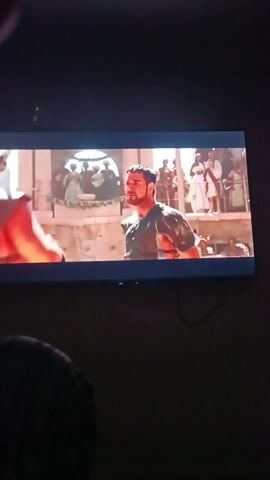 The video shows a screen displaying a scene from a movie. The scene appears to be an action sequence set in a desert-like environment. In the foreground, a man with short dark hair is seen wearing a dark sleeveless shirt. He has a determined expression on his face. Behind him, a large, brown, textured object, possibly a vehicle or a structure, is visible. In the background, there are glimpses of people in the distance and a red banner or cloth flying. The sky is a clear blue. The image quality suggests it's a recording of a screen, potentially from a phone or camera.