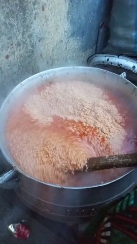 A large silver pot sits on a stove, filled with a reddish liquid and grains. A wooden stick is used to stir the mixture. Steam rises from the pot. The background shows a concrete wall, a seat, and a discarded can. The person stirring is wearing a colorful garment.