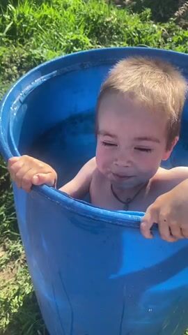 A young boy, with short brown hair, wearing a cross necklace, is immersed in a blue plastic barrel filled with water. He is smiling and looking directly at the camera. The barrel is sitting on a grassy area outdoors.  A wooden fence and other outdoor elements are visible in the background.