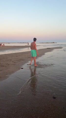 A person in turquoise swim trunks walks on a wet sandy beach near shallow water. They are walking away from the camera towards the sea. The sky is a gradient of pale blue and pink. In the distance, there are people on the beach and some breakwaters. The camera then pans to the sea where a person is visible in the water. Finally, the camera focuses on a person in a white skirt walking into the ocean with the sun setting in the background. The sky is a vibrant orange and pink.