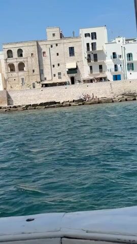 The video captures a sunny coastal scene from a low perspective, likely from a boat with a white railing visible at the bottom. Choppy, dark blue-green water occupies the foreground. Above the water, a light-colored stone wall forms the coastline, upon which approximately five to seven individuals are visible sitting or lounging. Behind the wall, several multi-story buildings line the shore under a clear, bright blue sky. A large, older stone building with arched windows is central, flanked by modern white buildings featuring balconies and blue window shutters. Soft music is playing in the background.