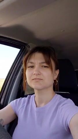 The video shows a woman in the driver's seat of a car. She is wearing a purple t-shirt and hoop earrings. Her hair is pulled back. She is looking to her left and speaking. The interior of the car is visible, including the dashboard and a portion of the passenger seat. The window is down, and a blurry outdoor landscape is visible outside. The lighting suggests it is daytime. The woman appears to be in the middle of a conversation or speaking to someone.