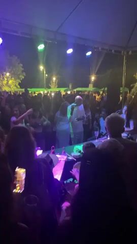 The video shows a live music performance at night, under a tent-like structure with stage lighting. A large crowd is visible, with many people dancing. A bald man in a white shirt is singing into a microphone on stage. Behind him, another person is playing percussion instruments. Tables with drinks are visible in the foreground, close to the stage. Several attendees are filming with their smartphones. The atmosphere is lively and festive.