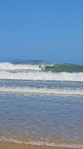 A surfer is riding a wave in a video. The video is shot at beach level with several rows of waves in the foreground. The person is dressed in a black wetsuit, and can be seen changing position and angling the surfboard on the face of the wave. The wave breaks at multiple points, and more waves appear behind it. The sky is blue with mountains visible in the distance. A single bird flies overhead.