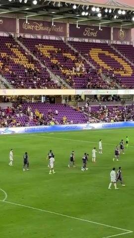 The video shows a wide shot of a soccer game taking place in a stadium. The stands are filled with spectators, most of whom are wearing purple. The field is green, and there are several players on the field wearing white and purple uniforms. A large screen is visible in the background, displaying game information. The sky above the stadium is a soft pink and orange, indicating it is either sunrise or sunset. The overall atmosphere appears to be one of excitement and engagement.