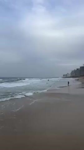 The video captures a beach scene on a cloudy day. The sandy beach stretches out towards the horizon, with gentle waves rolling onto the shore. Several people are scattered along the beach; some are wading in the water, while others appear to be surfing. In the background, a line of buildings can be seen along the coastline. The sky is overcast with gray clouds, lending a slightly muted tone to the overall scene.
