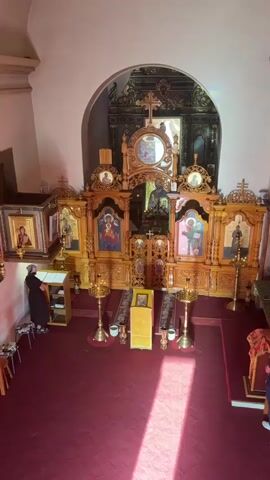 The video shows the interior of an Eastern Orthodox church, viewed from a high angle. In the foreground, a person in black religious attire is standing at a lectern, appearing to read. The focal point of the scene is an ornate, wooden iconostasis, which is a wall of icons and religious imagery separating the sanctuary from the nave. The iconostasis is richly carved and decorated, featuring several religious icons depicting figures in traditional Byzantine style. In front of the iconostasis, there are several golden candle stands (candlesticks) and a small, yellow-covered pedestal with an icon on top. The floor is covered with a red carpet, and sunlight streams in from an unseen window, creating bright stripes of light across the floor. Several other people are visible in the background, including two individuals standing near the right side of the frame, one of whom is looking towards the iconostasis. The overall impression is one of solemnity and religious devotion within a richly adorned ecclesiastical space.
