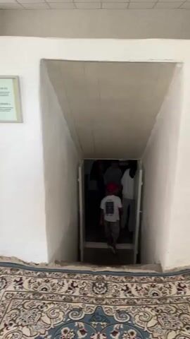 The video shows an interior scene with a downward sloping passageway leading into a dimly lit area. A few people, including children, are visible near the entrance of this passageway. In the foreground, two boys are standing, looking towards the passageway. The floor in the foreground is covered with a patterned rug. The walls are white and the ceiling of the passageway appears to be made of wood panels. The lighting is dim, particularly within the passageway.