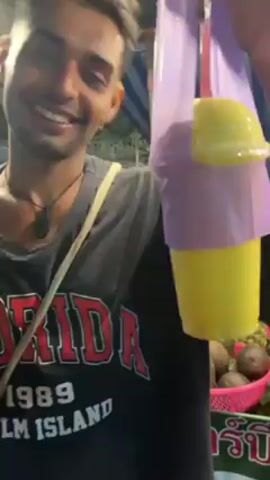 A person with tan skin, dark hair, and a small beard is standing outside and smiling. They are wearing a black t-shirt with "FLORIDA 1989 PALM ISLAND" in red and white letters. They have a small dark pendant necklace and a crossbody bag strap. In their right hand they hold a yellow beverage in a clear plastic cup, covered with a clear dome lid. The cup is wrapped in a purple plastic bag, and has a pink straw sticking out of the lid. To the person’s right, there is a pink plastic basket full of brown objects, and a green-and-white sign written in Thai. The location appears to be an outdoor market at night, with string lights overhead. The person turns to the side and waves in an underhand motion. Another person walks by in the background.