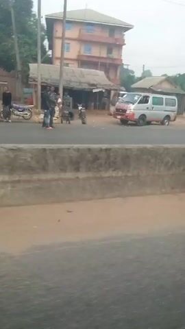 The video depicts a roadside scene in what appears to be a developing country. A van is parked on the right side of the road. Several people are visible, some standing near a collection of small shacks or shops. A man in a red shirt is sitting on a concrete barrier. A silver SUV passes by. The road is asphalt, and the environment appears dusty or hazy. Power lines are visible overhead.