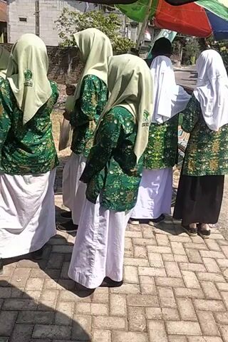 The video displays a line of people, predominantly young women, dressed in Islamic attire. They are standing on a brick-paved surface under what appears to be daylight, with some vegetation and buildings visible in the background. The individuals are wearing headscarves (hijabs) and long garments. Some are adorned with green and white patterned vests or outer garments. The camera angle is rotated, making the scene appear vertical. The people seem to be standing in a procession or formation. There are a few individuals who are less clearly visible at the top of the frame, possibly further down the line or at a distance. The scene suggests an outdoor event, potentially a school activity or religious gathering.