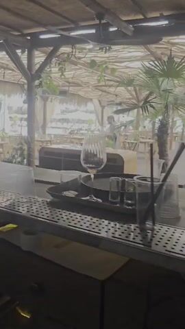 The video shows a bar area with a thatched roof. In the foreground, a bar counter is visible with various bar tools and ingredients, including glasses, shakers, and cut fruit in stainless steel containers. In the background, there is an outdoor seating area with tables and chairs, some of which are covered with white canopies. Palm trees and other greenery are present, suggesting a tropical or beachside setting. A person, possibly a staff member, is seen in the background walking through the seating area. The overall atmosphere appears relaxed and possibly pre-opening or during a quiet period.