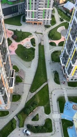 The video shows an aerial view of a modern residential complex. The focus is on the courtyard and the surrounding high-rise apartment buildings. The courtyard features green spaces, paved pathways, benches, and playground equipment. Several people are visible walking in the courtyard and on the sidewalks. The architecture of the buildings is characterized by their height, modern design, and use of light colors with green and yellow accents. The sky is partly cloudy, suggesting it is daytime. The surrounding environment appears to be a suburban or semi-urban area with other buildings and greenery visible in the distance. The camera is positioned high up, likely from another building, providing a bird's-eye perspective of the scene. The video is brief and shows a static view of the complex with no significant movement or action.