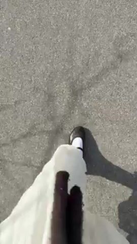 The video shows a person walking along an asphalt path. The person is wearing black Mary Jane shoes with white socks and a long white dress. The path is bordered by grass and trees on one side and a field on the other. In the distance, there are houses. The sky is blue with some clouds. The video appears to be shot on a sunny day.