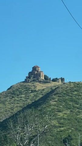A video shows a view of a stone building with a domed roof situated on a green, tree-covered hill under a clear blue sky. A tall, white pole stands vertically in the foreground on the right side of the frame, obscuring part of the view. A thin dark wire runs diagonally across the top right corner of the frame. The camera appears to be moving slightly, causing a subtle shift in perspective of the landscape.