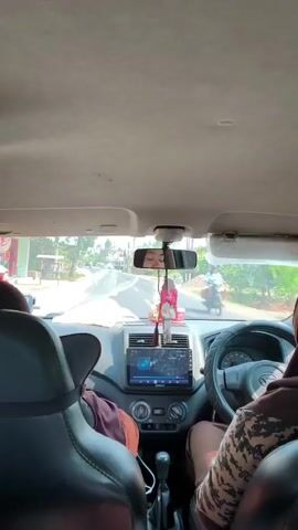 The video is taken from the back seat of a car, looking forward through the front windshield. The driver, a person wearing a hijab, is visible in the driver's seat, with their hands on the steering wheel. A passenger is in the front passenger seat, also wearing a hijab. The car is moving along a road, which appears to be in a rural or suburban area, with trees and buildings visible on either side. The sky is bright and appears to be daytime. The dashboard of the car is visible, including a touchscreen infotainment system. A decorative item, possibly an air freshener or a charm, is hanging from the rearview mirror. The camera appears to be handheld and is somewhat shaky.