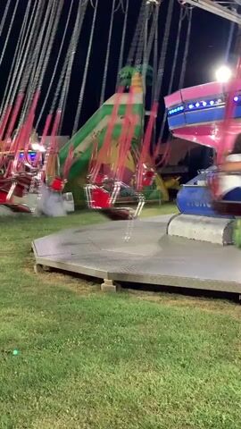 A swing ride at a nighttime fair is in motion, with several empty seats and some occupied by people. The ride consists of chairs suspended by chains from a rotating central mechanism, which appears to be made of metal with pink and blue accents. The ride is surrounded by a grassy area and in the background, there is a colorful inflatable structure resembling a tropical scene with palm trees. The lighting is artificial and bright, illuminating the ride and the surrounding area. There is also a small amount of smoke or fog being emitted from the base of the ride. The people in the chairs are blurred due to motion, but some appear to be leaning back and enjoying the ride. One person is visible in a pink outfit and another in white pants and a top. A person in a dark t-shirt is seen near the central mechanism. The overall atmosphere is one of amusement and excitement.