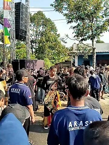 The video shows an outdoor event with a crowd of people. In the center of the frame, two individuals are dressed in elaborate costumes, one of whom appears to be wearing a headdress and ornate clothing. There are other people in the crowd, some wearing t-shirts with logos that include the word "ARASI." A large speaker system is visible on the left side of the frame. The event seems to be taking place in a street or open area with buildings in the background. The atmosphere appears festive.