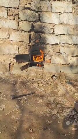 A small fire burns in a brick fireplace embedded in a light-colored brick wall. The flames are orange and yellow, reaching upwards, and the firebox has a metal door that is partially open. The ground in front of the fireplace is dirt and scattered debris, including twigs and leaves. A dark metal tool is lying on the ground to the left of the fireplace. The wall behind the fireplace shows signs of soot from previous fires. The scene appears to be outdoors in sunlight.