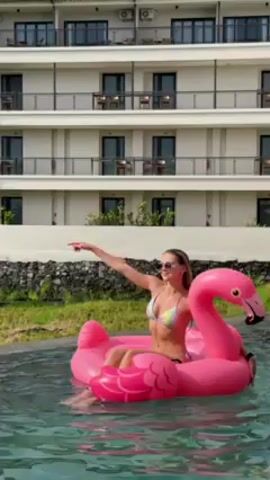 A young woman is floating on a pink inflatable flamingo in a pool. She is wearing a bikini and sunglasses, and raises one arm up in the air. She then puts her arms down and relaxes. The pool is in front of a white multi-story building with black doors and railings on the balconies. There is green grass and some stones between the pool and the building. The water in the pool is a bright, clear blue color. Music is playing in the background.
