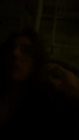 The video appears to show two people in low light, possibly at night. The person in the foreground has long hair.  The second person, who is closer to the right side of the frame, is visible from the eyes up.  The background is dark but suggests the presence of a wall or structure with possible window. Music plays.