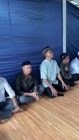 The video shows a group of men sitting on the floor in a room. The men are wearing traditional Muslim clothing and some are wearing skullcaps. There is a blue curtain in the background and a wooden floor. The men are facing a table with two men sitting behind it. There are also some women in the foreground, also sitting on the floor. The men behind the table appear to be speaking or leading a prayer.
