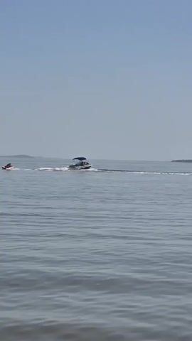The video shows a sunny day on a body of water. Initially, a boat tows someone on an inflatable behind it. The boat moves towards a small island in the distance. Next, the shot changes to three women standing in the water. They appear to be conversing and looking out at the water.