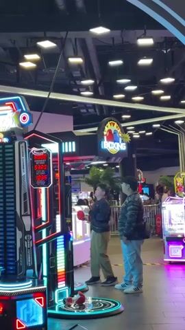 The video shows an indoor arcade with rows of game machines. A person wearing tan pants and a black jacket is standing in front of a "Storm Hammer" punching game, preparing to punch the pad. To the right of the person, another person wearing blue pants and a camouflage jacket is standing, looking at the game. In the background, other people can be seen walking around. The lighting is bright and the ceiling is high with exposed pipes. The arcade is clean and well-maintained. A sign for a "Boxing Game" is visible above the machines.