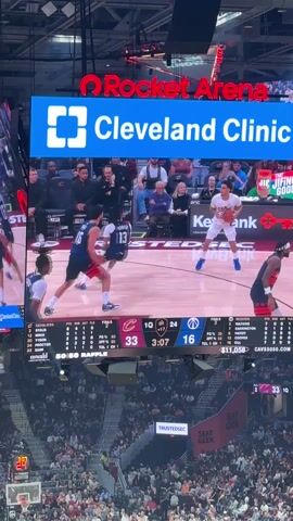 The video shows a basketball game in progress at Rocket Arena, sponsored by the Cleveland Clinic. The scoreboard shows the Cleveland Cavaliers leading with 33 points against their opponents who have 16 points. It’s the first quarter with 3 minutes and 3 seconds left. Players are on the court, and a player in a white jersey is seen shooting a basketball. The stands are filled with spectators watching the game. Banners displaying advertisements from Seat Geek, Trustedsac, and KeyBank are visible around the arena.