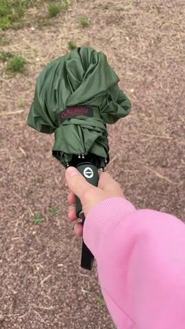 The video shows a first-person perspective of someone holding a closed green umbrella in their right hand. The background is a gravelly path with sparse dry grass and some green vegetation. The person is wearing a pink hoodie. The umbrella is held by its handle, which appears to be dark green. The umbrella is folded and strapped closed with a dark green fabric loop. The person's hand grips the handle, and the thumb is visible near the umbrella's mechanism. The video is short and seems to capture a moment of holding or preparing to use the umbrella.