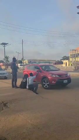 The video shows three people and a red Toyota RAV4 on a street corner in daylight. A man in uniform stands near the front of the vehicle, holding a long, dark object.  A second man in a white shirt and dark pants sits on the curb with his back to the car, pointing towards it. Another person wearing dark clothing kneels on the ground near the sitting man. The red RAV4 has its front passenger side tire partially on the sidewalk. Behind the vehicle is a road with other vehicles, including cars and a motorcycle. In the background, a yellow and white two-story building is visible, as well as utility poles and street lights. The sky is partly cloudy.