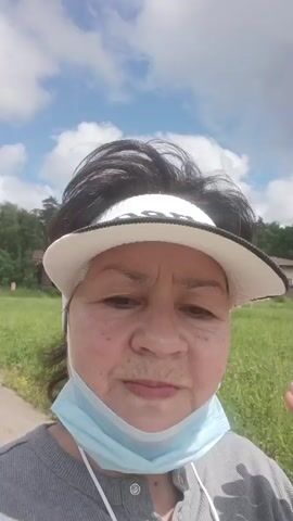 The video begins with a close-up selfie of a woman wearing a white visor with black trim, a grey shirt, and a light blue face mask. She has dark hair pulled back and is outdoors on a sunny day with a partly cloudy sky. In the background, there is a grassy field with some trees and buildings, including a light-colored house. The woman is looking at the camera and then turns her head to the side, speaking.

The video then pans out to show a view of a paved path or road leading into the distance. On either side of the path, there are grassy areas and scattered buildings, some appearing to be wooden structures. The sky is still partly cloudy, suggesting it is daytime. The overall setting appears to be a rural or semi-rural area. There is no discernible text in the video. The woman's facial expressions suggest she is engaged in conversation, possibly describing the weather or her surroundings. The presence of the face mask implies that the video was recorded during a time when mask-wearing was recommended or required. The location context indicates this is likely in Russia.