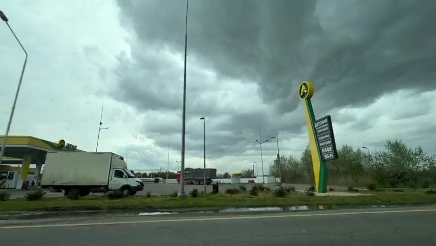 The video provides a point-of-view shot from a vehicle driving along a road under a heavily overcast and dark sky, suggesting an impending storm or a gloomy day. On the right side of the road, a gas station with a distinctive yellow and green color scheme and a matching logo (possibly an "A") comes into view. Multiple fuel pumps are visible under its canopy. A white box truck is parked near the gas station, and a red truck can be seen further in the background. A red circular "No Entry" sign is prominently displayed near the gas station's entrance. As the vehicle continues, it passes a stretch of undeveloped land or a field, followed by a two-story red brick house. Numerous streetlights and utility poles carrying overhead power lines are visible along the roadside throughout the video. The scene depicts a roadside environment, likely in the Tliustenkhabl' area of Russia, characterized by infrastructure like the gas station and power lines, alongside residential and undeveloped plots.